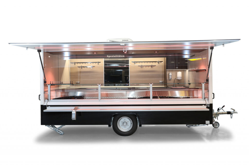 Verkaufsanhänger mit einer Verkaufskühltheke von 4,5m. Verbaut ist ein Stilles Kühlsystem. Die Arbeitsfläche an der Verkaufstheke ist mit einer beheizten Körperschutzkante ausgestattet. An der Verkaufsrückwand befinden sich 2 Hakenschienen aus Edelstahl und eine Werbetafel  mit Glasschriftfeld. Unterhalb der Hakenschienen befinden sich Unterschränke und das Hygienepaket.