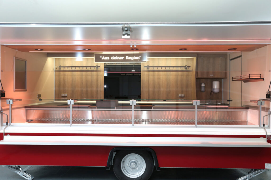 moderner Verkaufsanhänger-Kühltheke mit 740 mm Thekenauslage und Doppelkühlfach für 4 E-Kisten-Hakenschienen, Schalttafel 400 Volt, Glaswerbetafel, senkrechter Thekenaufsatz und Körperschutzkante mit integrierter Heizung